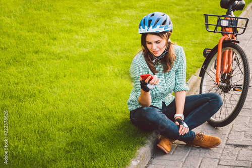 Man and city rolling bicycle, environmentally friendly transport. Beautiful young caucasian woman worker sitting resting on the grass uses a red mobile phone near an orange bicycle with a coryne