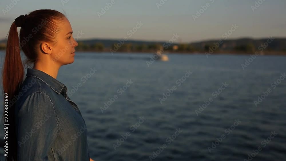 young beautiful Caucasian middle-aged woman with red hair in a blue denim dress stands near the sea on the waterfront in the evening at sunset against the blue sky. young beautiful Caucasian middle-aged woman with red hair in a blue denim dress stands near the sea on the waterfront in the evening at sunset against the blue sky.