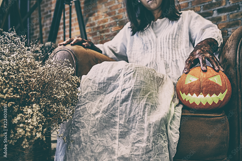 Halloween festival concept. Ghost girl in the blood with white dress holding the halloween pumpkin and sitting on old leather sofa.