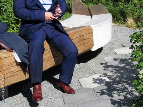 A man in a dark blue business suit and brown leather shoes sitting on a bench in the park with a smartphone in his hands. Concept for busy man, businessman or official