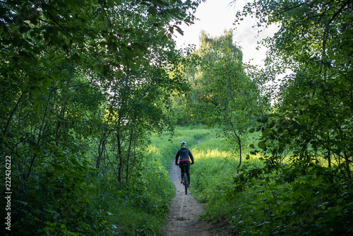 Photo from back of woman bicyclist wearing helmet riding along road in woods