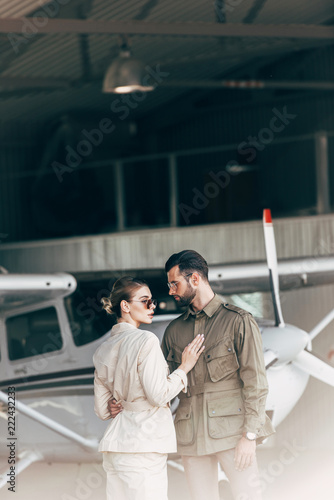  fashionable young couple in stylish jackets standing in hangar with airplane