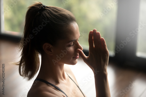 Fotografie Young attractive woman practicing yoga, doing Alternate Nostril Breathing exercise, nadi shodhana pranayama pose, working out, indoor close up, yoga studio, side view
