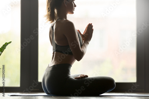 Young sporty attractive woman practicing yoga, doing Padmasana exercise, Lotus pose with namaste gesture, working out wearing sportswear grey pants and top, indoor yoga studio. Body close up view