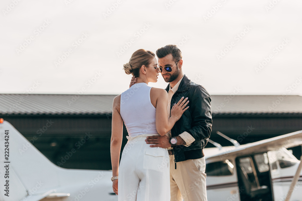 stylish man in leather jacket and sunglasses embracing attractive girlfriend near plane