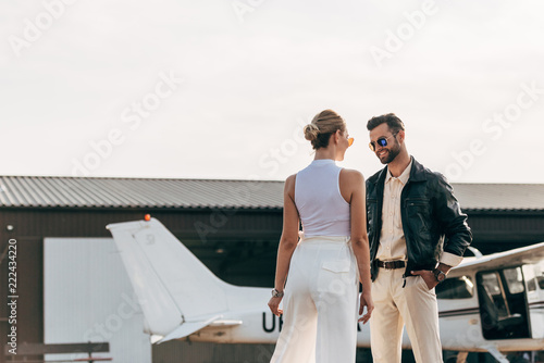 rear view of stylish woman talking to smiling boyfriend in leather jacket and sunglasses near plane
