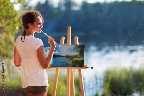 Young pretty woman artist draws paints a picture of a lake on open plain air outdoors