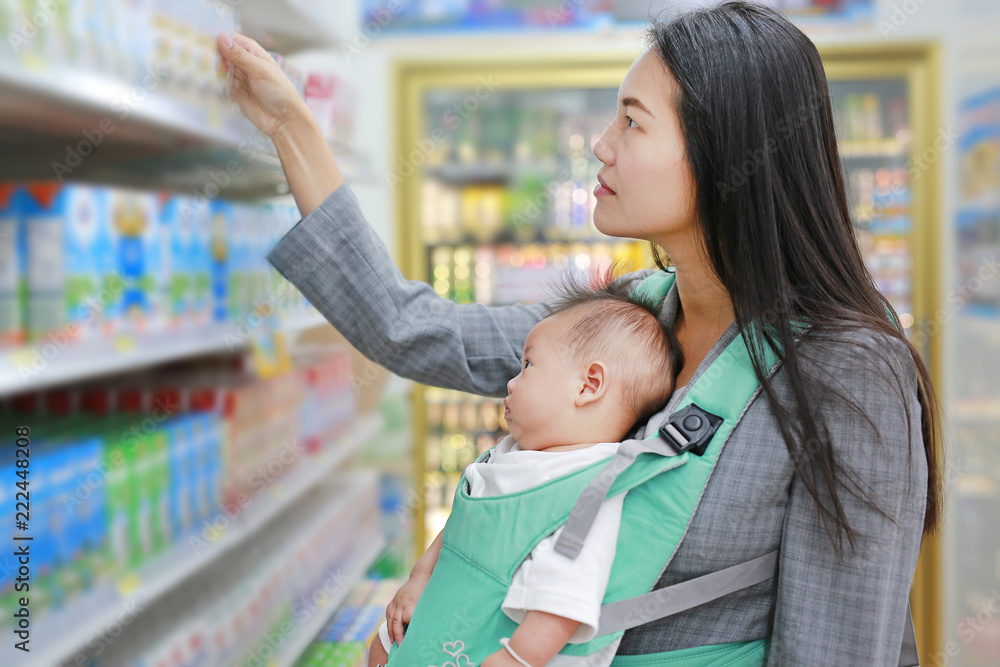 Young business woman choosing breakfast in Convenience Store while carrying her baby boy by hipseat.