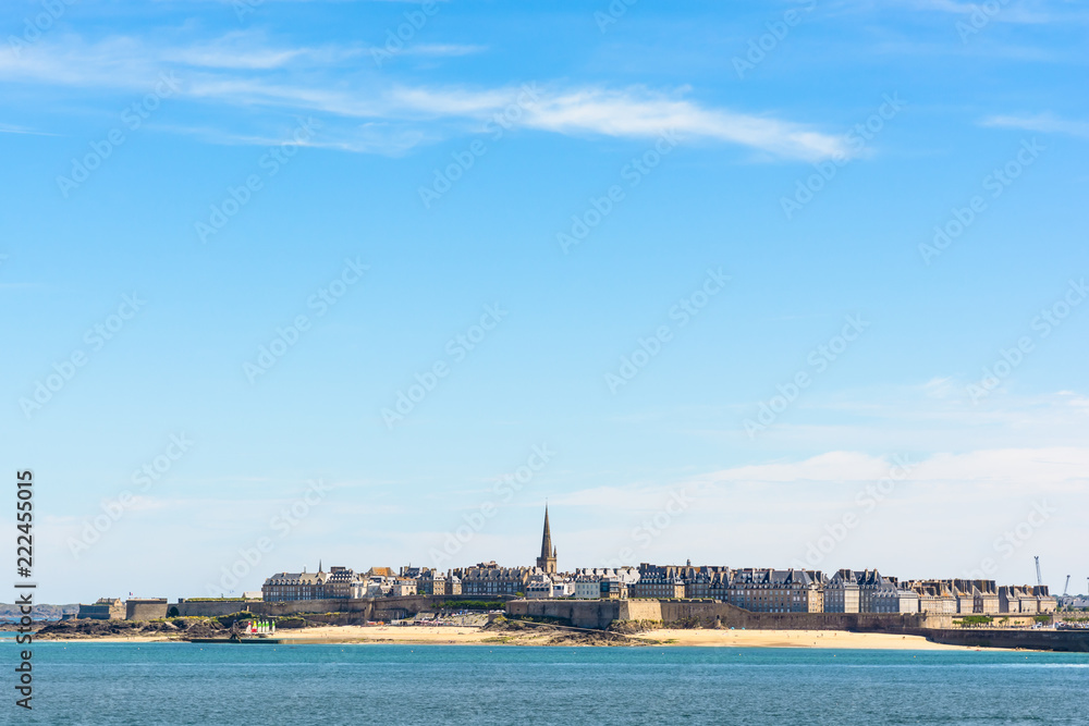 General view of the walled city of Saint-Malo in Brittany, France, with the steeple of the cathedral protruding above the buildings behind the wall under a large portion of blue sky.