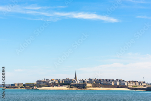 General view of the walled city of Saint-Malo in Brittany, France, with the steeple of the cathedral protruding above the buildings behind the wall under a large portion of blue sky.