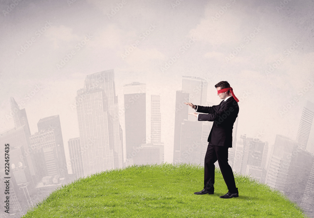 Young blindfolded businessman steps on a a patch of grass with a cloudy city in the background