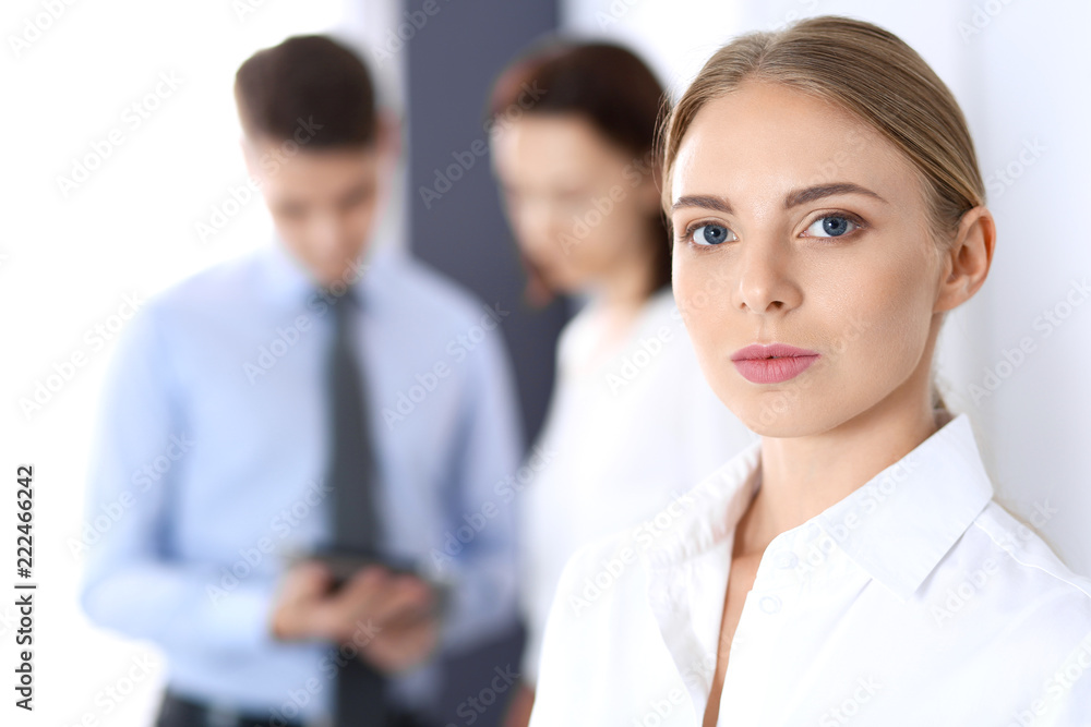 Beautiful blonde businesswoman standing straight in a brightly lit office at the background of colleagues or partners. Business concept