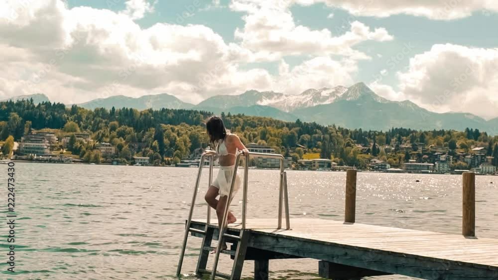 Young woman in swimwear climbs down the stars on a dock in Worthersee lake, mountain view windy hair in slow-motion Young woman in swimwear climbs down the stars on a dock in Worthersee lake, mountain view windy hair in slow-motion