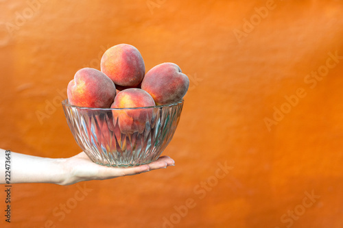 Profile side view photo of young woman hand hold full glass crystal transparent bowl of big beautiful ripe mellow peaches isolated on vivid orange background with copy space for text photo