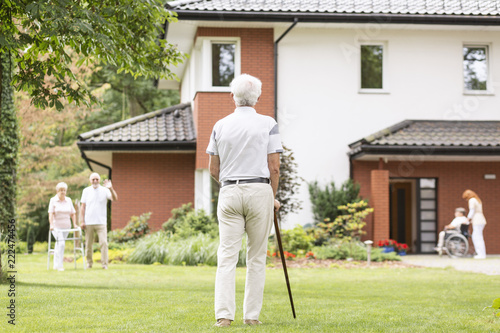 A back of a gray-haired senior man with a cane looking at his fellow mates outside of a luxury nursing home.