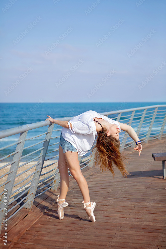 Naklejka premium Young attractive ballerina training on sea coast wearing casual. Fine art ballet concept. Surreal dancing. Slim girl dance on the ocean beach. Active and healthy women