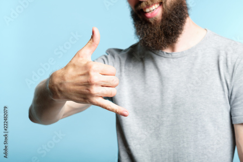 bearded man showing phone call gesture or shaka sign with hand. cropped shot of a male torso on blue background. casual hipster in grey t-shirt. photo