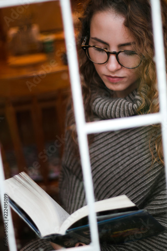 Beautiful, warm portrait through window with drops of water of young adult reading on a rainy day
