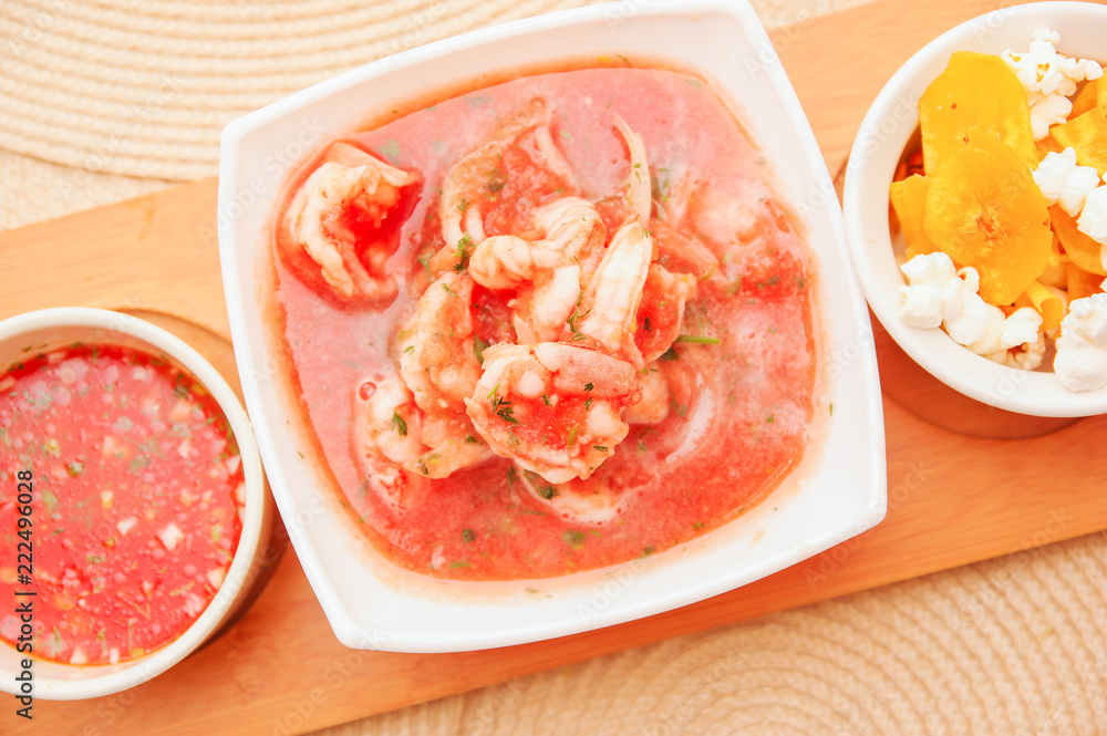 Above view of delicious shrimp cebiche in rectangular white bowl served with chifles and red spicy sauce over a wooden structure in table background