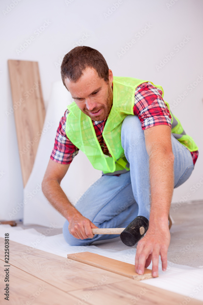 Smiling young man wearing reflective safety vest installing laminate flooring in new apartment or house, holding a hammer. Home improvement and renovation concept Smiling young man wearing reflective safety vest installing laminate flooring in new apartment or house, holding a hammer. Home improvement and renovation concept