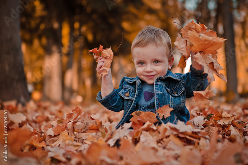 Little happy boy in blue jacket is playing with leaves at golden autumn park background