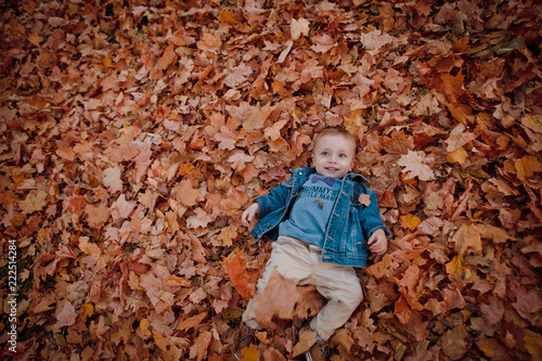 Little happy boy in blue jacket is playing with leaves at golden autumn park background