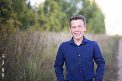 portrait of a smiling guy in a blue shirt against a background of trees and a dirt road to the field © artem
