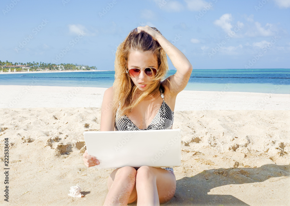 A woman sitting on the sand at the beach working on her computer with a shocked expression A woman sitting on the sand at the beach working on her computer with a shocked expression