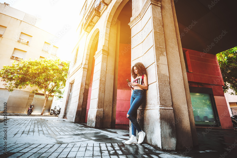 Fototapeta premium A svelte young charming caucasian female with beautiful curly hair is leaning against the arch column on the street and typing a message to her friend using the smartphone, sunny day in Barcelona