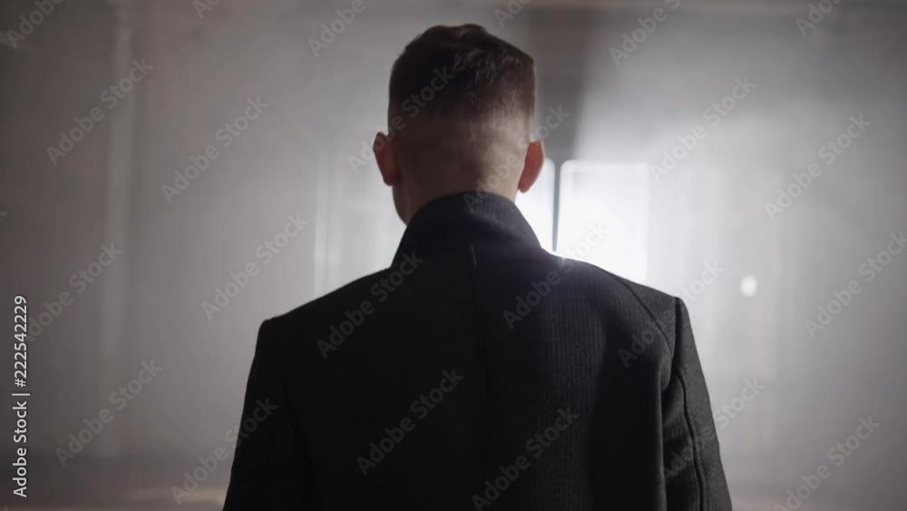 Short haired man wearing long grey jacket with straight collar is walking in darken empty hall towards bright lighting coming out of open windows Short haired man wearing long grey jacket with straight collar is walking in darken empty hall towards bright lighting coming out of open windows