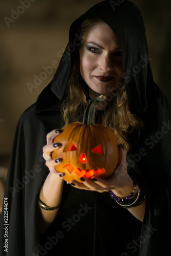 Beautiful witch in black mantle with a pumpkin face on Halloween