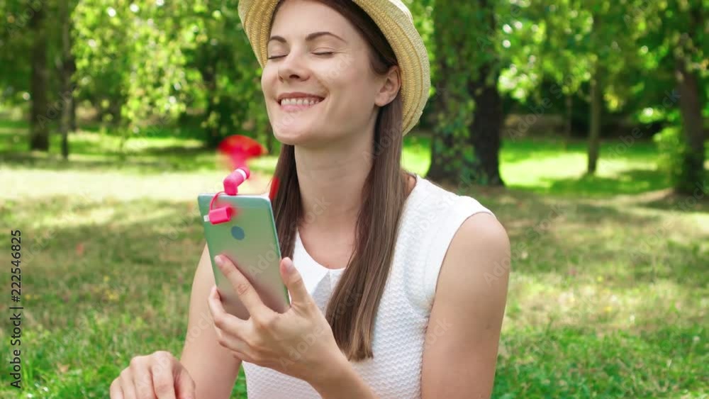 Female student sitting on grass on college campus using pink portable ventilator attached to mobile phone during heat. Tourist in hat cooling down with small fan working from smartphone in public park Female student sitting on grass on college campus using pink portable ventilator attached to mobile phone during heat. Tourist in hat cooling down with small fan working from smartphone in public park
