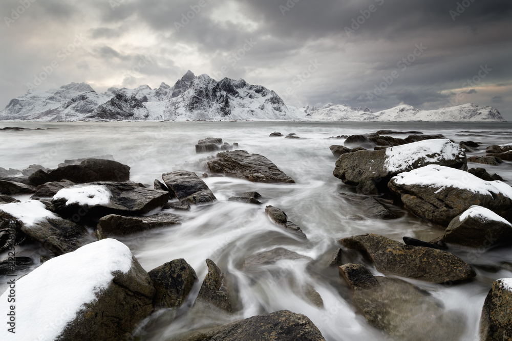 Coastal landscape in winter with water movement between big stones, in the background a mountain range with snow, high contrast sky - Location: Norway, Lofoten