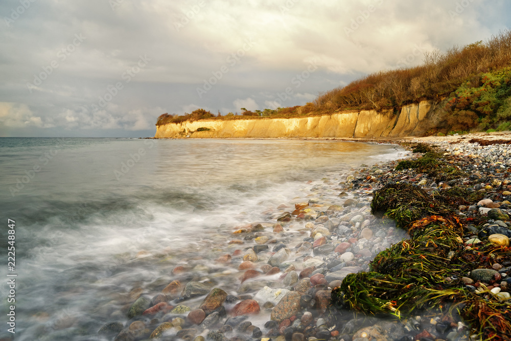 Obraz premium Wide beach with seaweed and stones in the evening light, overlooking the water and the steep bank, water movement in long exposure - Location: Germany, Rügen Island
