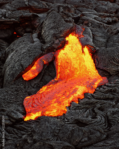 A lava flow emerges from a rock column and pours into a black volcanic landscape, the hot lava shows up in yellow and red shades - Location: Hawaii, Big Island, volcano "Kilauea"