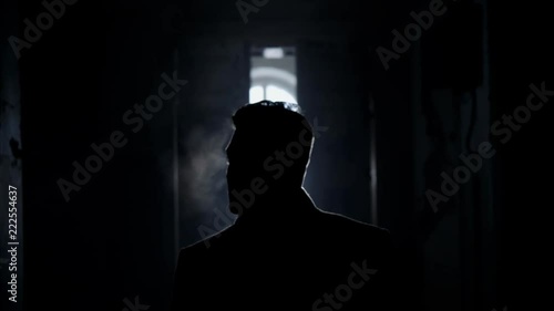 Silhouette of mature short haired man dressed in long coat with straight collar walking along dark corridor towards empty bright room with white dusty walls and big arched windows.