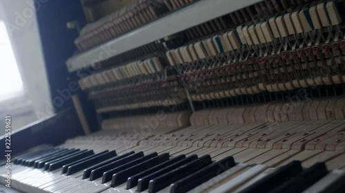 Classic wooden brown piano with broken body frame and dusty keys having revealed inside hammers and strings is depressively abandoned in empty light hall surrounded by white walls and large windows.