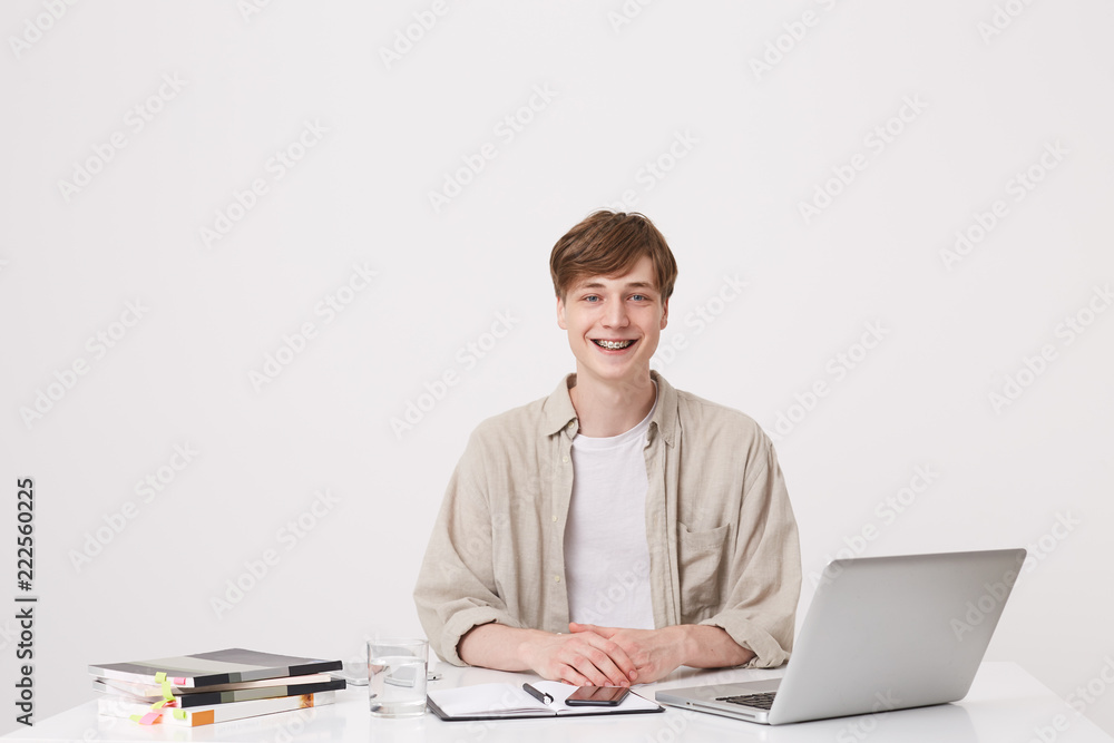 Closeup of cheerful young male student with braces wears beige shirt study using laptop computer and notebooks sitting at the table isolated over white background Closeup of cheerful young male student with braces wears beige shirt study using laptop computer and notebooks sitting at the table isolated over white background