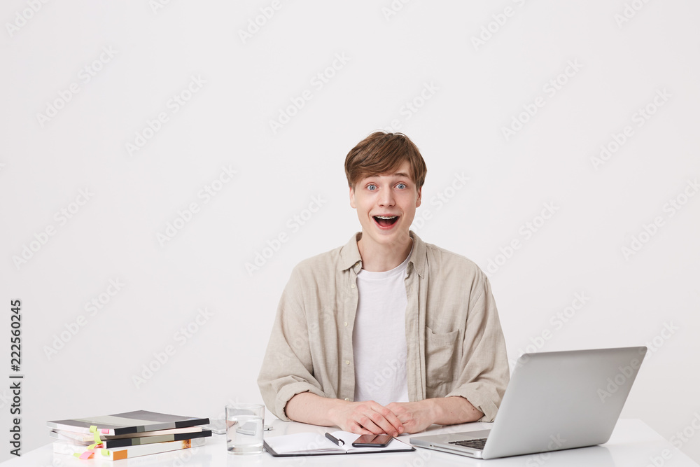 Closeup of surprised young man student with braces wears beige shirt feels impressed and using laptop computer and notebooks at the table isolated over white background Closeup of surprised young man student with braces wears beige shirt feels impressed and using laptop computer and notebooks at the table isolated over white background