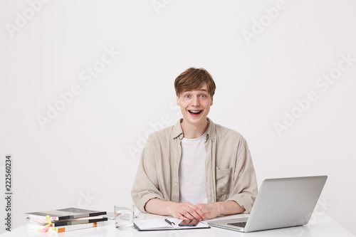 Portrait of amazed young man student wears beige shirt looks surprised and study at the table with laptop computer and notebooks isolated over white background