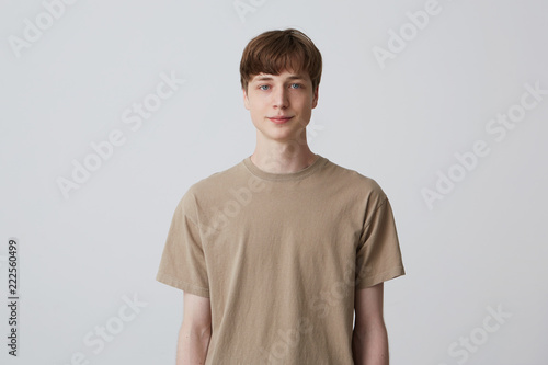 Portrait of handsome attractive young man with short haircut and blue eyes wears beige t shirt standing and smiling isolated over white background Looks directly in camera