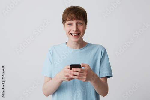 Closeup of happy young man student with short haicut and braces on teeth wears blue t shirt holding and using cell phone isolated over white background