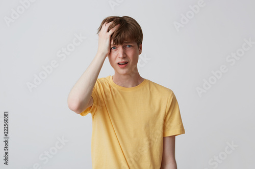 Portrait of desperate unhappy young man with hand on head and braces on teeth in yellow t shirt looks sad and having a headache isolated over white background