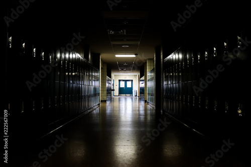 Straight view of darkly lit hallway with bright light at end and lockers covering the walls on either side 