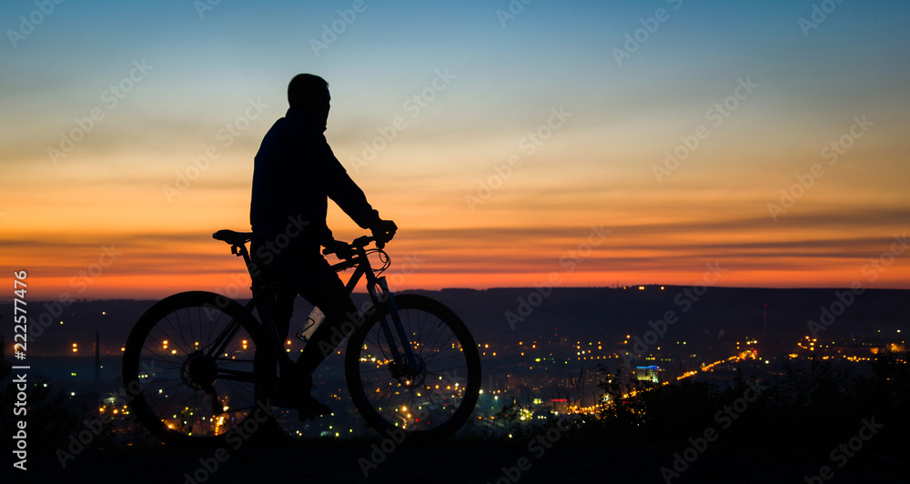 Obraz premium A young cyclist rests on the top of the mountain near the city, at sunset.