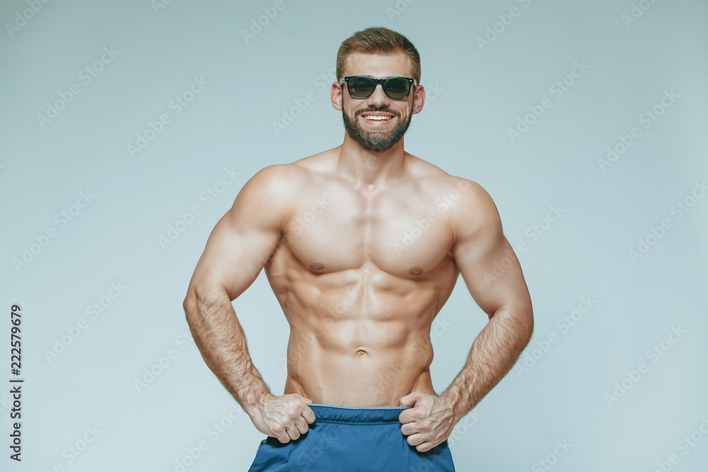 .bodybuilder posing. Beautiful sporty guy male power. Fitness muscled in blue shorts and sunglasses. on isolated grey background. Man with muscular torso. Strong Athletic Man Fitness Model Torso .bodybuilder posing. Beautiful sporty guy male power. Fitness muscled in blue shorts and sunglasses. on isolated grey background. Man with muscular torso. Strong Athletic Man Fitness Model Torso