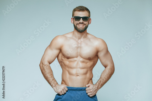 .bodybuilder posing. Beautiful sporty guy male power. Fitness muscled in blue shorts and sunglasses. on isolated grey background. Man with muscular torso. Strong Athletic Man Fitness Model Torso