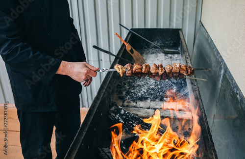 Man in black uniform is frying meat pork on the grill. Cook is making shish kebab for summer party. Shashlik on wooden panels and open fire.