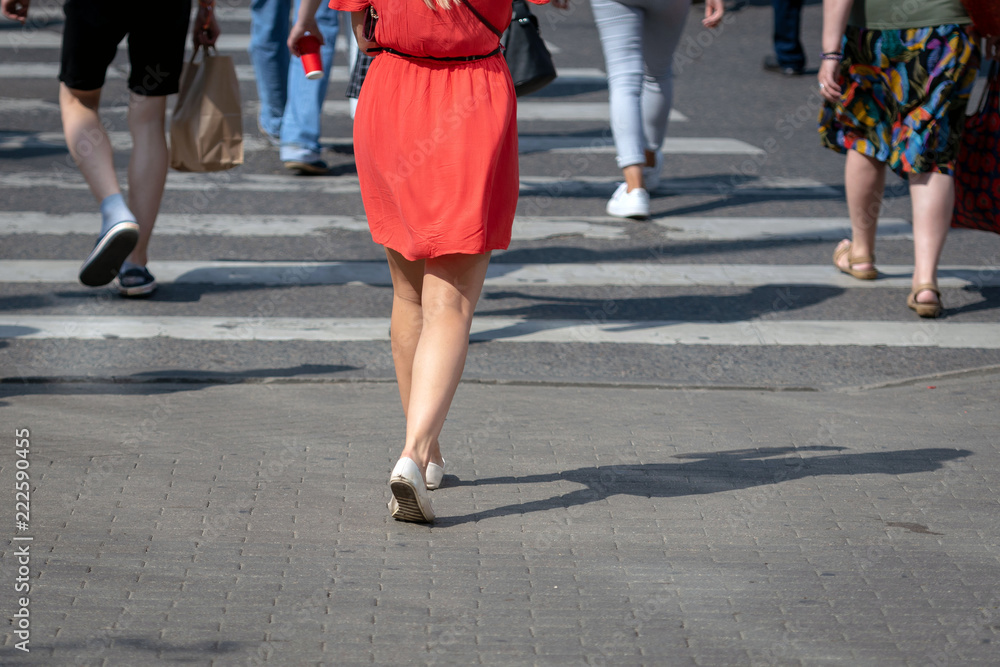 On a sunny day, pedestrians cross the street through a pedestrian crossing. There are pedestrian shadows on the street. View from the back.