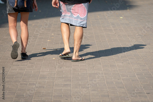 On a sunny day, a couple of people walk along the sidewalk. The human shadows are visible on the sidewalk. View from the back.